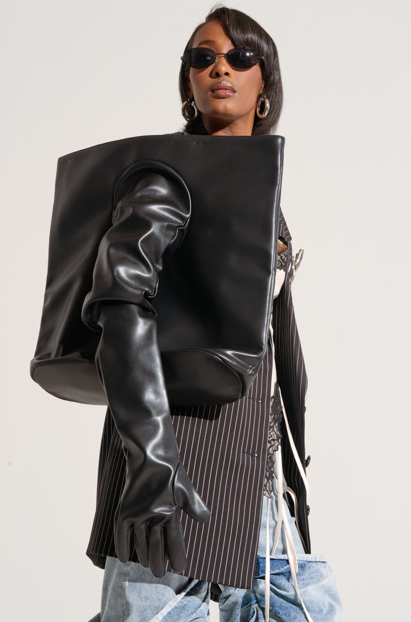 A person wearing dark sunglasses, a pinstripe blazer, and a statement bag paired with unique oversized black leather accessories, including the POST ME GLOVE PURSE and a boxy shoulder piece. The attire is avant-garde with light blue baggy jeans and layered clothing elements.