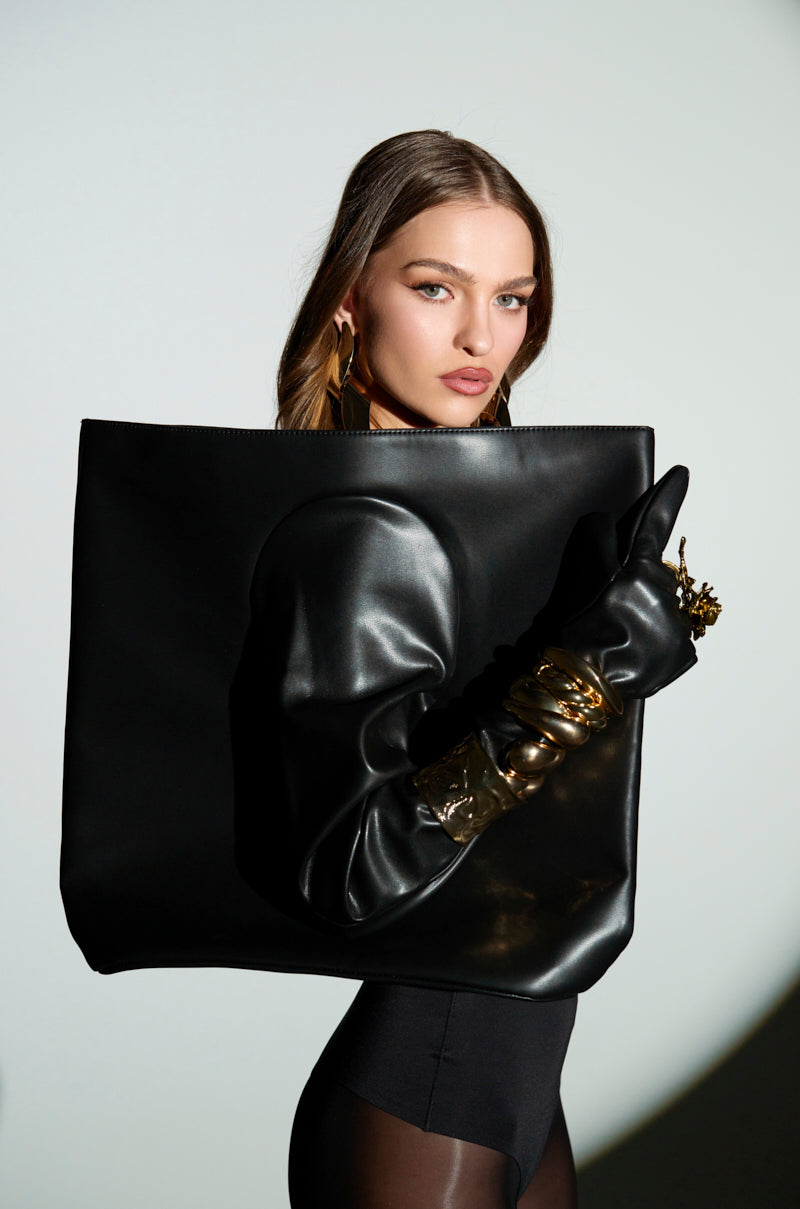A woman poses confidently with the POST ME GLOVE PURSE in front of her. She sports statement gold earrings, a black top, gold bangles, and black gloves against a light background.