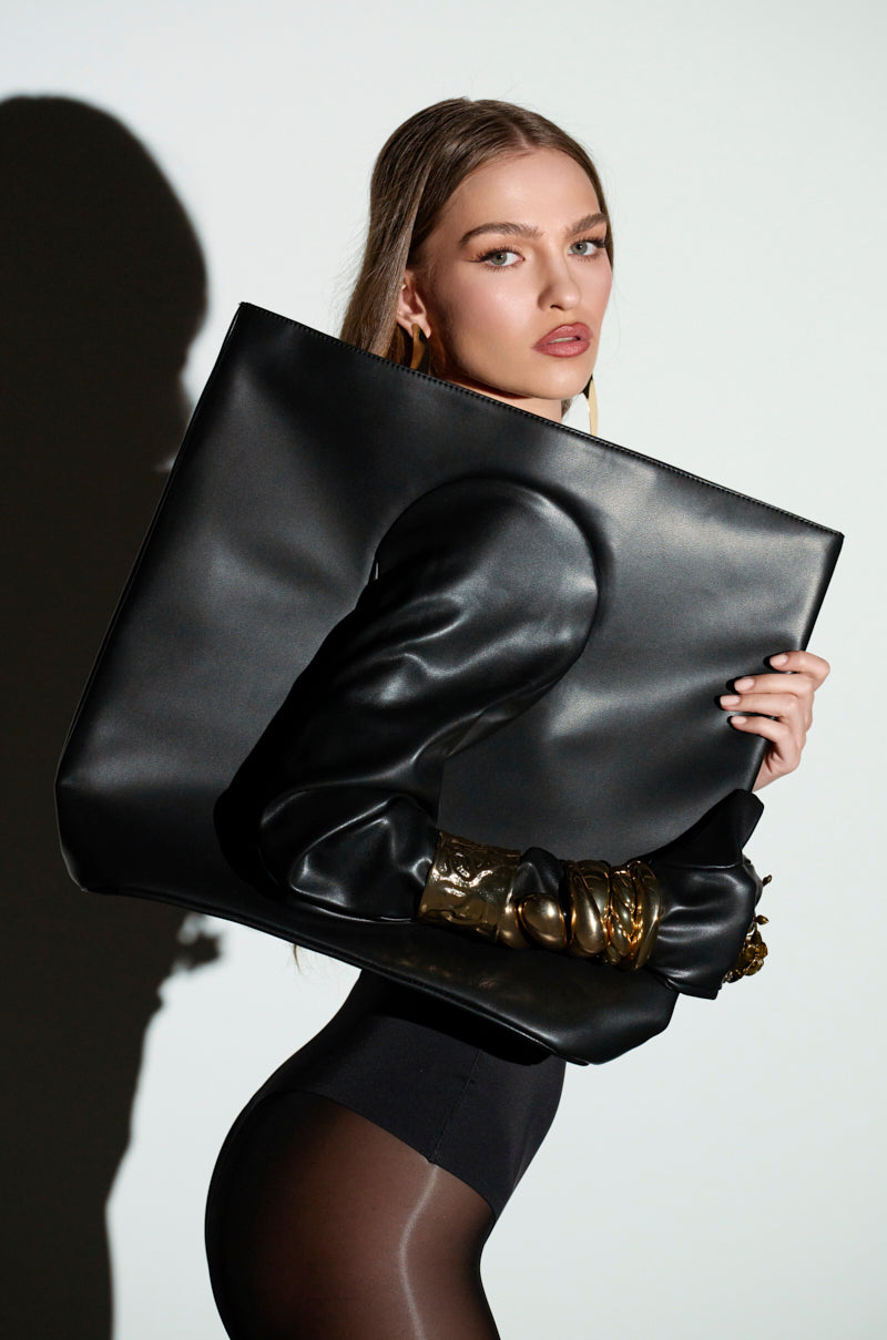 A person with long hair poses against a light background holding a POST ME GLOVE PURSE by AKIRA Label. They sport a black outfit with sheer sleeves and gold accessories, creating an interesting silhouette effect as their shadow intertwines with that of the Glove Purse.