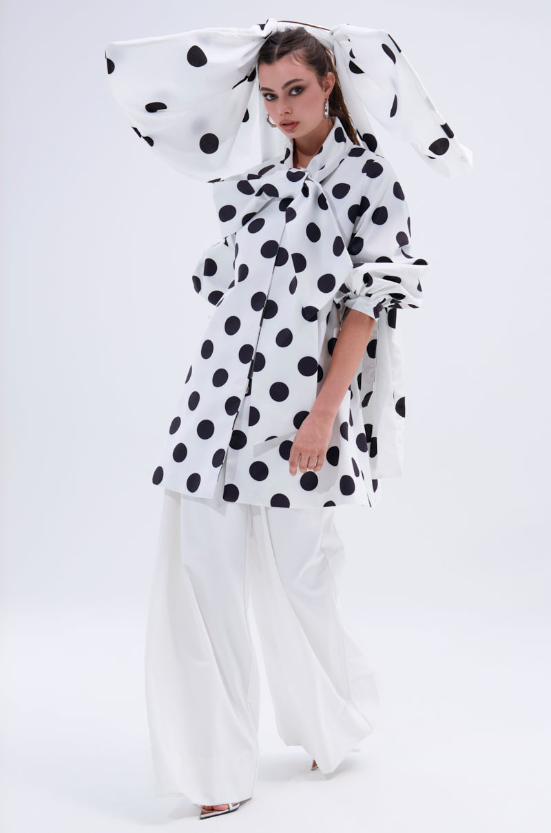 A woman models the AMALIA TIE NECK TRENCH IN POLKA DOT, a striking white coat with oversized black spots, styled with a large bow headpiece, loose polka-dot top, and wide-legged white pants against a plain light background.