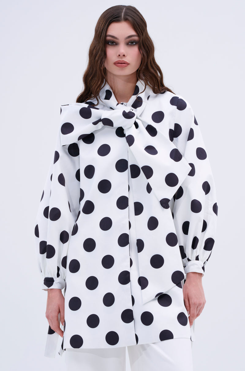 A woman with long wavy hair models the AMALIA TIE NECK TRENCH IN POLKA DOT, featuring balloon sleeves and an oversized neck bow. She stands against a plain light background, showcasing this chic statement piece.