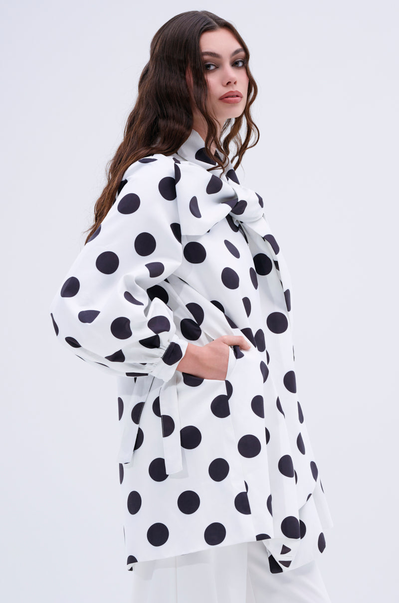 A woman with long wavy hair stands in a relaxed pose, one hand in her pocket, wearing the AMALIA TIE NECK TRENCH IN POLKA DOT—white with bold black spots and a statement bow—against a simple light background.
