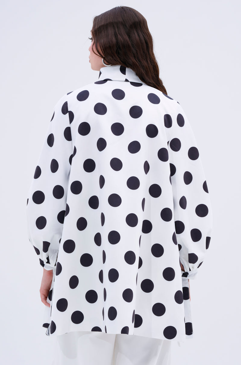 A person with long brown hair is seen from the back against a plain light background, wearing the AMALIA TIE NECK TRENCH IN POLKA DOT—a loose-fitting statement trench with bold black spots that stands out elegantly.