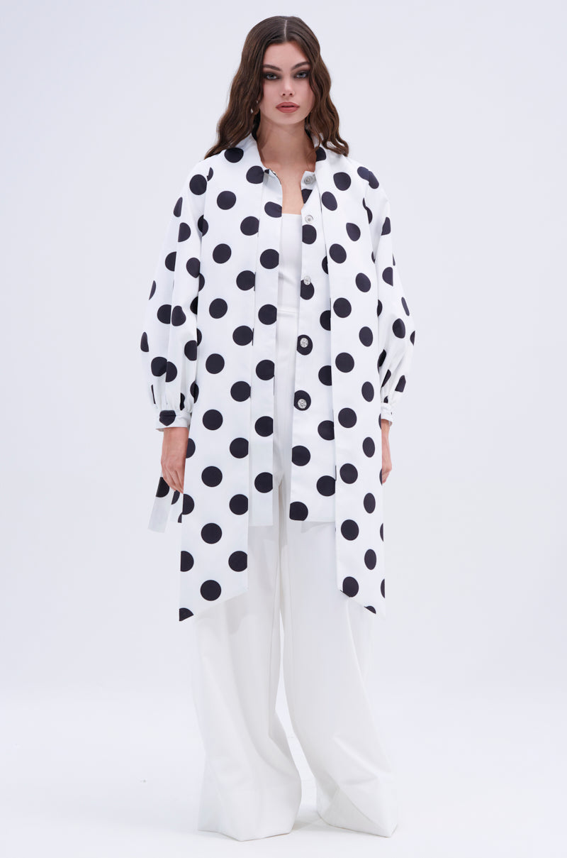 A woman with long, wavy brown hair and a neutral expression wears the AMALIA TIE NECK TRENCH IN POLKA DOT—white with bold black spots—over wide-leg pants against a plain background, making the statement trench the focal point of her look.