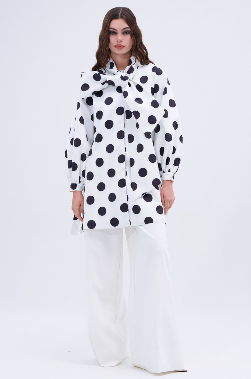 A woman wears the AMALIA TIE NECK TRENCH IN POLKA DOT, featuring a big neck bow, voluminous sleeves, and white wide-leg pants, set against a plain light background.