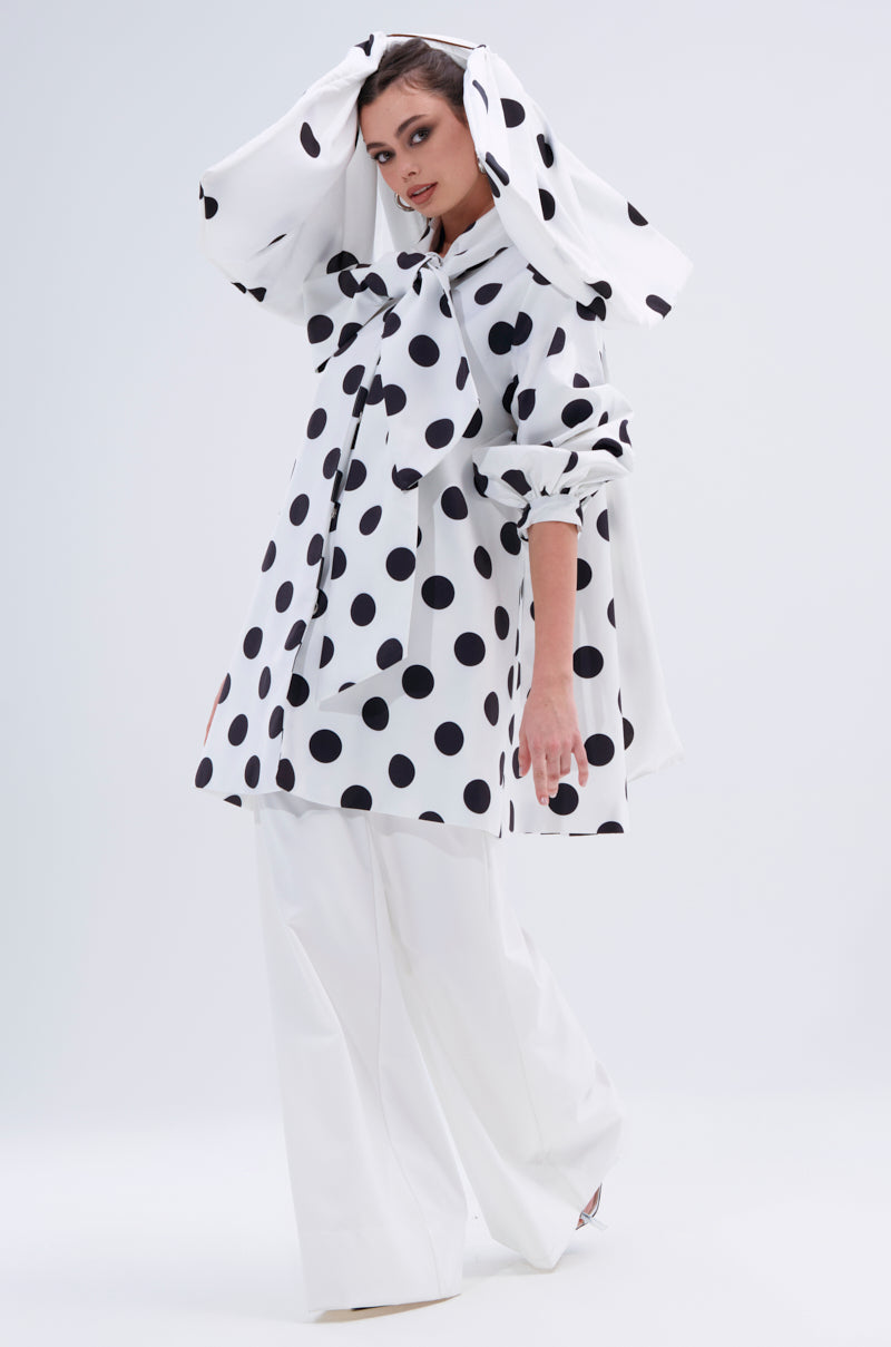 A person models the AMALIA TIE NECK TRENCH IN POLKA DOT, a bold hooded coat with large black polka dots, paired with wide-legged white pants against a simple light background.