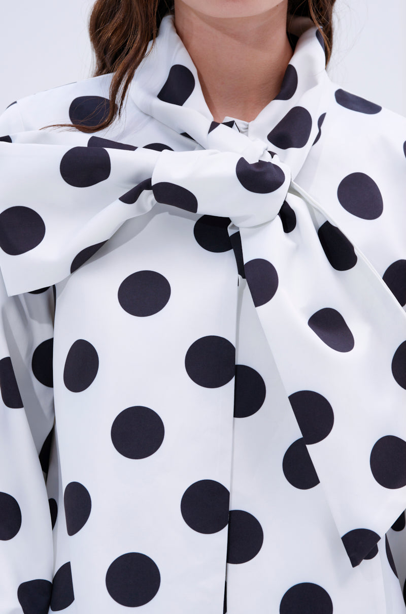 Wearing the AMALIA TIE NECK TRENCH IN POLKA DOT, a person showcases its bold white fabric with large black polka dots and a dramatic tie-neck bow. The person's face is partially out of frame.