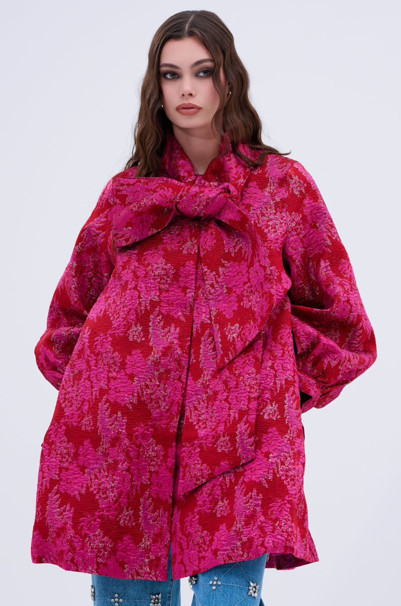 A woman with wavy brown hair models the AMALIA TIE NECK TRENCH, a vibrant pink and red brocade coat with a statement bow collar, standing against a plain light background.
