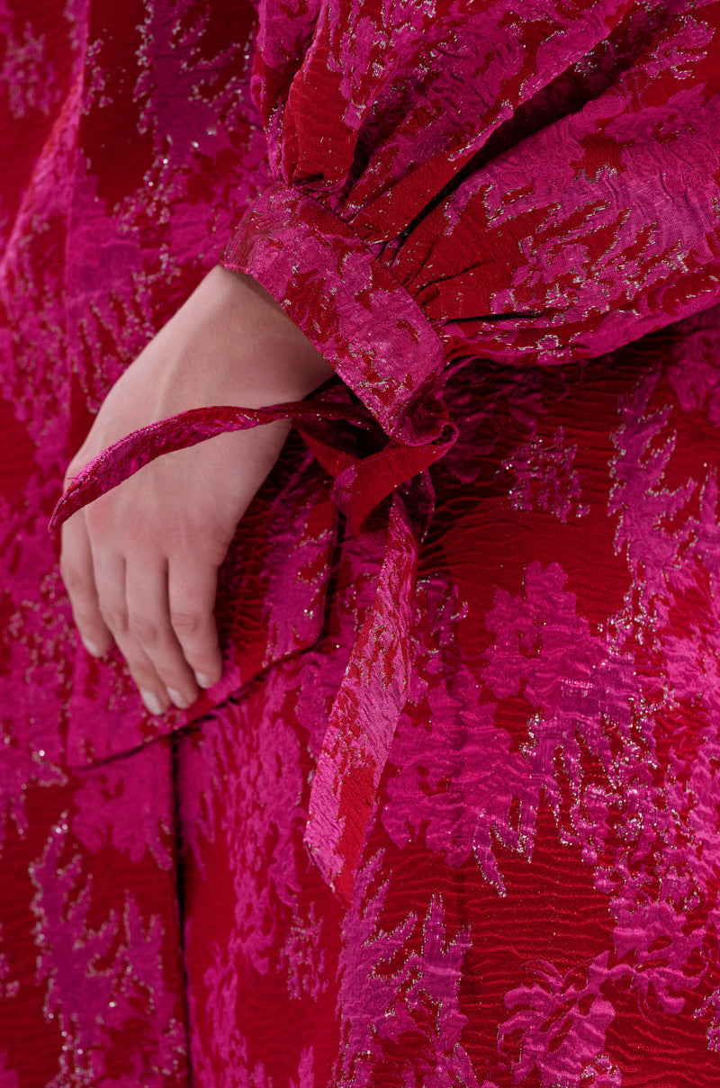 A close-up of a hand resting on the hip, wearing the AMALIA TIE NECK TRENCH—a red and pink brocade floral garment featuring a tied fabric bow on the sleeve.
