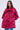 A woman with wavy brown hair models the AMALIA TIE NECK TRENCH, a vibrant pink and red brocade coat with a statement bow collar, standing against a plain light background.