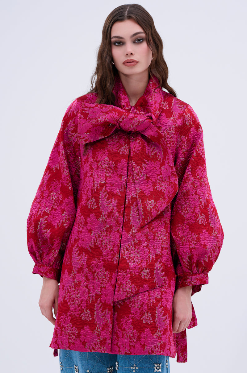 A woman models the AMALIA TIE NECK TRENCH, a vibrant long-sleeve coat with voluminous sleeves, a large bow at the collar, and a striking red and pink brocade pattern, set against a plain light background.