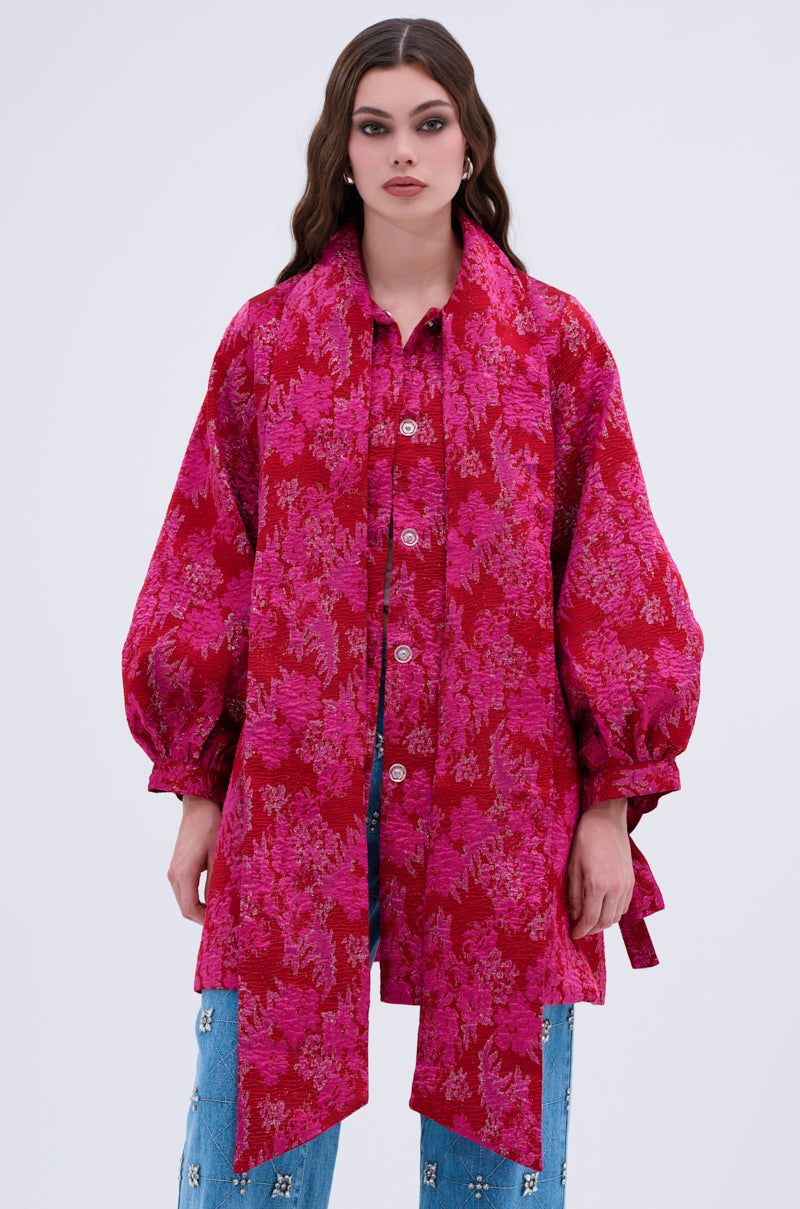 A woman with long wavy brown hair wears the AMALIA TIE NECK TRENCH, a loose bright pink brocade button-up with balloon sleeves and bow tie detail, paired with light blue jeans against a plain light background.