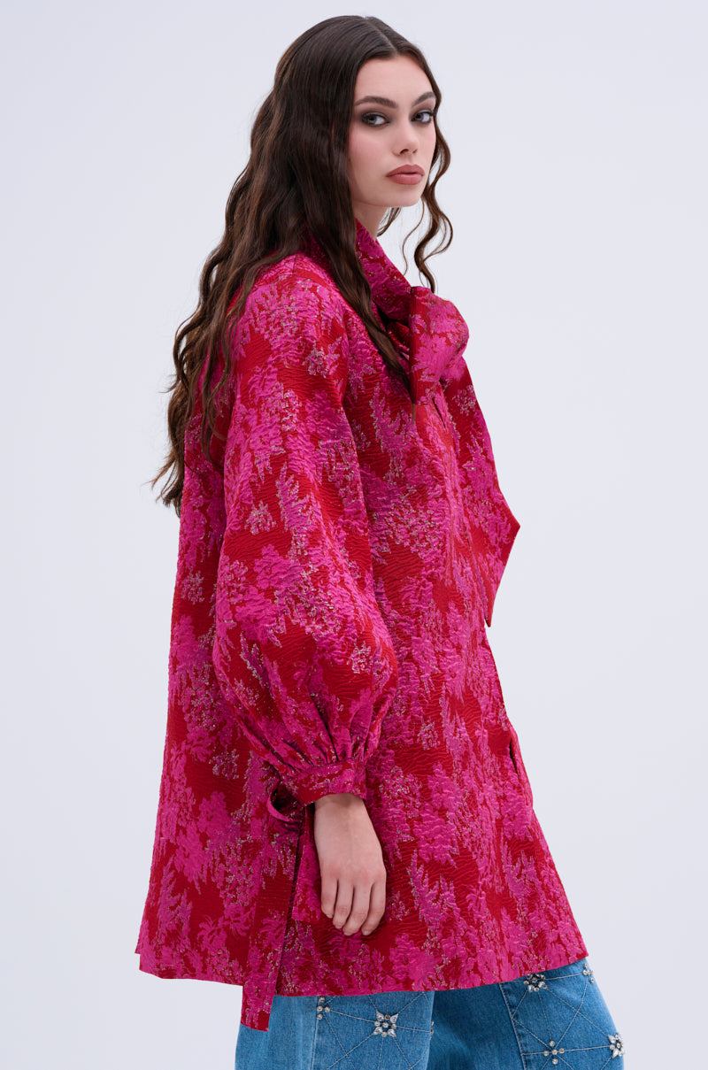 A woman with long, wavy brown hair wears the AMALIA TIE NECK TRENCH—a loose-fit, long-sleeved pink and red floral blouse with a brocade pattern and neck bow—paired with star-embellished blue jeans against a plain light background.