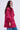 A woman with long, wavy brown hair wears the AMALIA TIE NECK TRENCH—a loose-fit, long-sleeved pink and red floral blouse with a brocade pattern and neck bow—paired with star-embellished blue jeans against a plain light background.