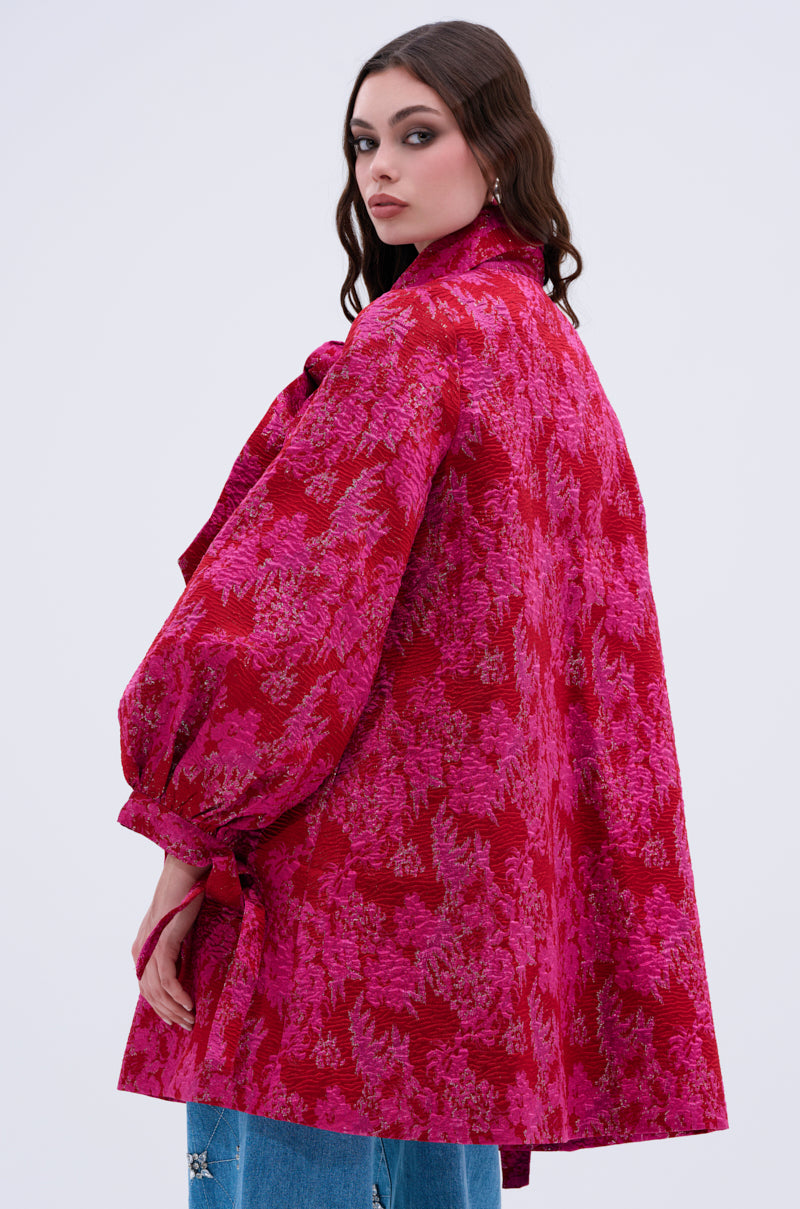 A woman with long brown hair wears the AMALIA TIE NECK TRENCH, a loose-fitting bright pink and red floral brocade coat with puffy sleeves and bow details, paired with blue jeans, standing against a plain light background.