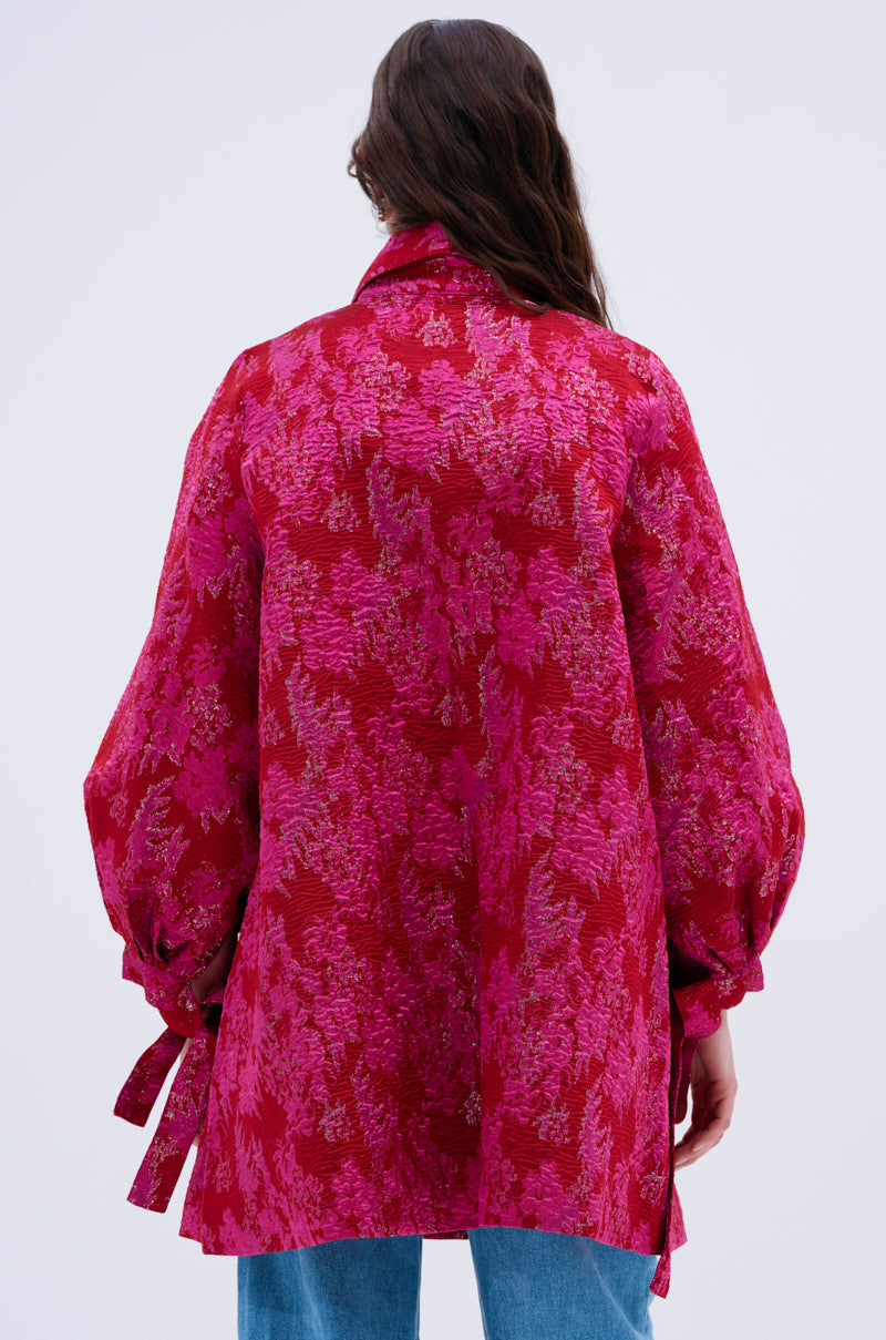 A person with long brown hair, seen from behind, wears the AMALIA TIE NECK TRENCH—a loose-fitting, bright pink and red floral brocade blouse with long sleeves, tie wrist details, and blue jeans.