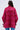 A person with long brown hair, seen from behind, wears the AMALIA TIE NECK TRENCH—a loose-fitting, bright pink and red floral brocade blouse with long sleeves, tie wrist details, and blue jeans.