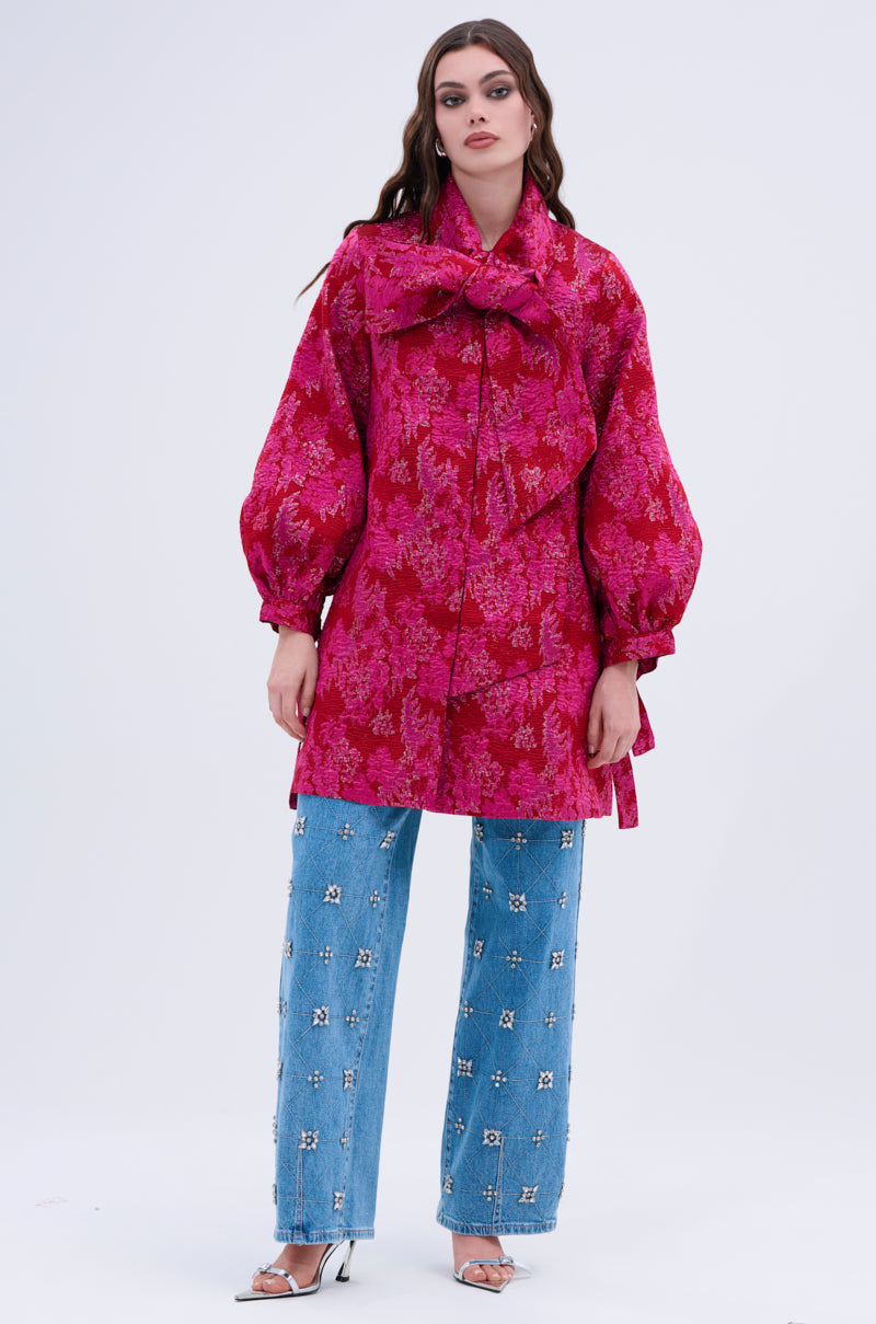 A woman poses against a plain background wearing the AMALIA TIE NECK TRENCH—a bright pink brocade long blouse with a large neck bow—paired with loose blue jeans featuring white embellishments and silver open-toe heels.