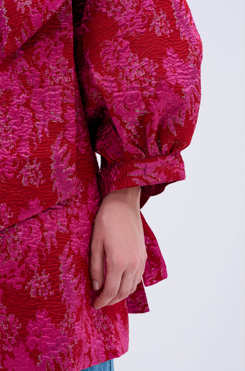 A person’s arm and hand rest by their side, wearing the AMALIA TIE NECK TRENCH—a textured red and pink brocade-patterned garment with puffy sleeves—against a light, plain background.