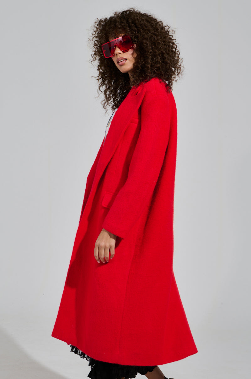 A person with curly hair is wearing large red sunglasses and the PHOEBE OVERSIZED TRENCH, made from a vibrant wool blend. They are standing sideways against a plain background.