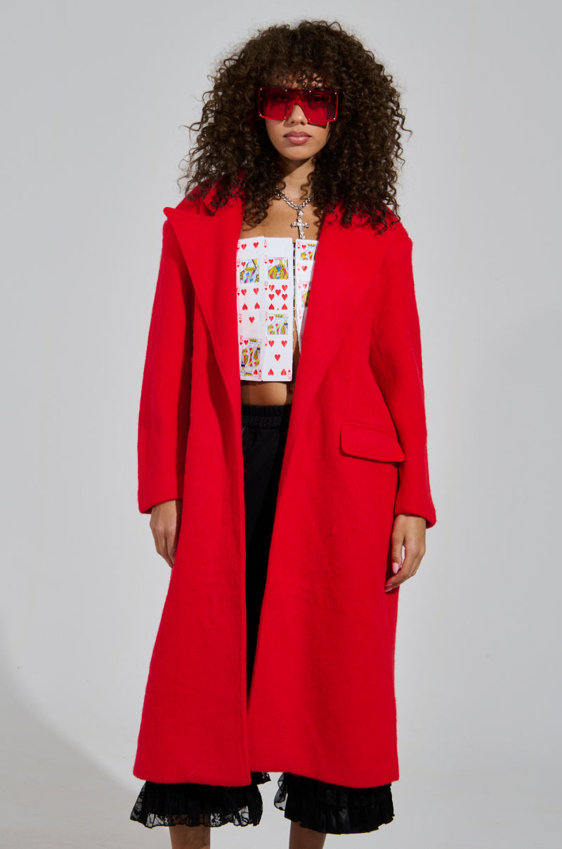 A person with curly hair wears oversized red sunglasses, a bright red PHOEBE OVERSIZED TRENCH featuring a longline design with a single visible pocket, a patterned crop top, and black pants. The background is plain and light-colored.