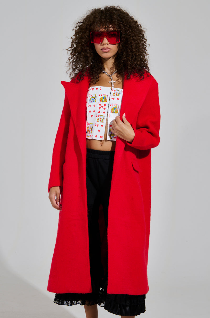 A person with curly hair is wearing oversized red sunglasses and the PHOEBE OVERSIZED TRENCH over a colorful patterned top and black pants. They are posing against a plain background.