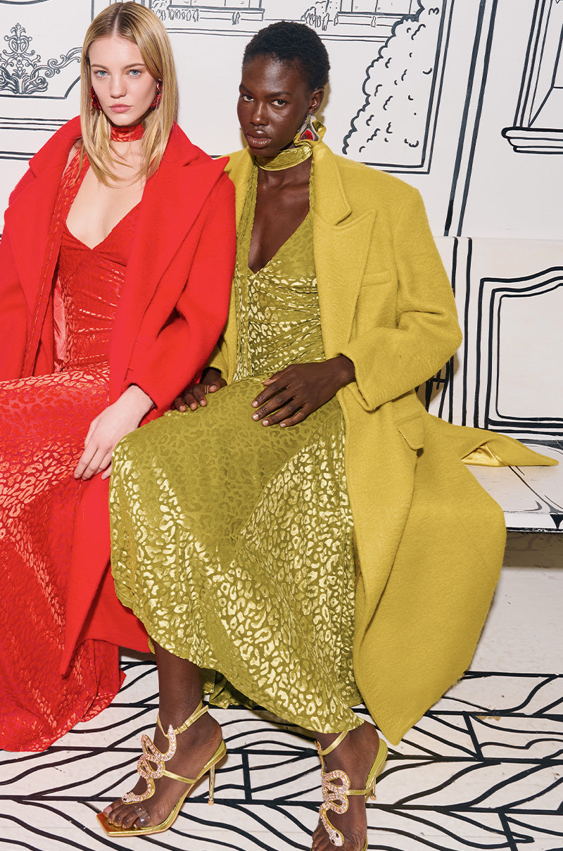 Two women sit stylishly on a black and white illustrated bench. One wears a red dress with a matching coat, while the other dons a PHOEBE OVERSIZED TRENCH IN GREEN over a patterned dress. Both have styled hair and colorful sandals, creating a fashion-forward ensemble.