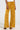 The shot captures the lower half of an individual donning Dripping in Gold crushed velvet trousers in a high-rise mustard yellow with a slight flare, complemented by brown heeled sandals. The background features a neutral, light color.