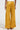 A person wearing the DRIPPING IN GOLD CRUSHED VELVET TROUSER is positioned against a light background, showcasing the back. These high-rise trousers have a wide-leg design and gracefully drape to the floor, partially concealing the person's sandals.