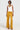 A person wearing a white sleeveless top, a layered necklace, and the DRIPPING IN GOLD CRUSHED VELVET TROUSER stands confidently against a plain background. Sporting sunglasses and wide-leg silhouette pants, they pose with their left hand on their hip.