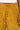 A close-up of a person wearing the DRIPPING IN GOLD CRUSHED VELVET TROUSER, featuring a wide-leg design in mustard yellow with a shiny, textured appearance. The person's hand rests near the side seam, and a white top is partially visible.