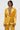 Wearing the DRIPPING IN GOLD CRUSHED VELVET BLAZER from the AKIRA Label, complemented by an intricate beaded necklace, a person stands against a plain background, exuding confidence while gazing at the camera.