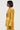 A person with a bob haircut stands in profile, adorned in gold, wearing the DRIPPING IN GOLD CRUSHED VELVET BLAZER by AKIRA Label, which features a textured golden-yellow design with an oversized collar. They have a serious expression against a plain background.
