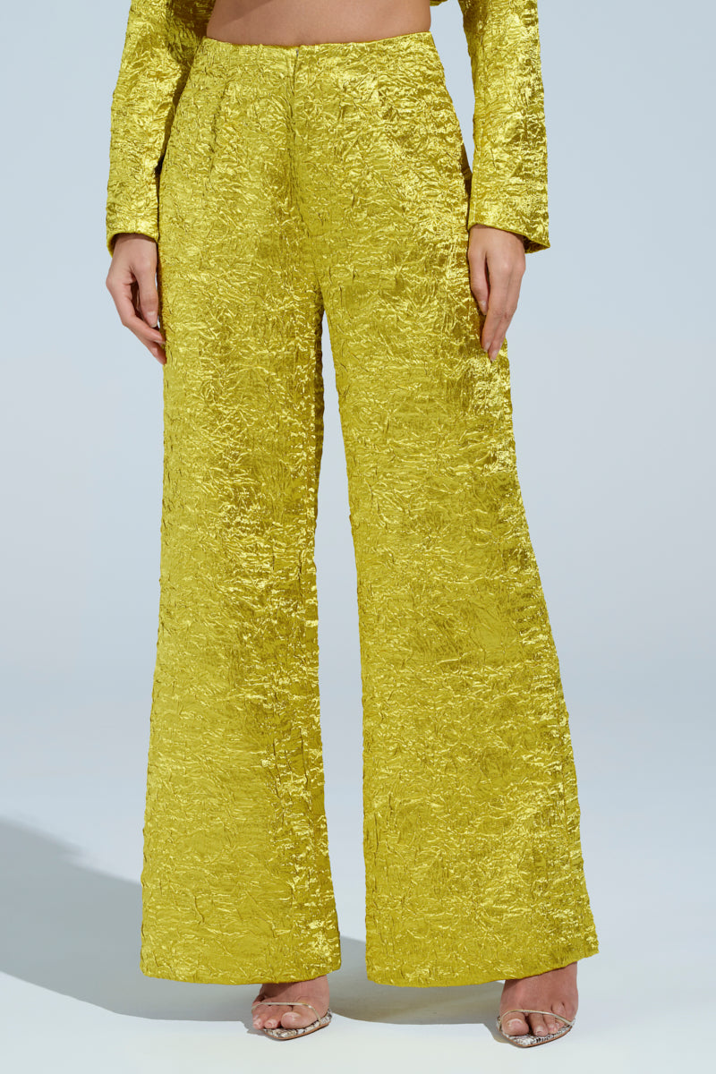 A person in the EXPENSIVE TO BE ME TROUSER, textured bright yellow wide-leg pants paired with a matching long-sleeve crop top. The high-rise fit flatters their silhouette, and the crinkled fabric adds depth. Posed on tiptoe in clear heels against a plain backdrop, they radiate effortless elegance.