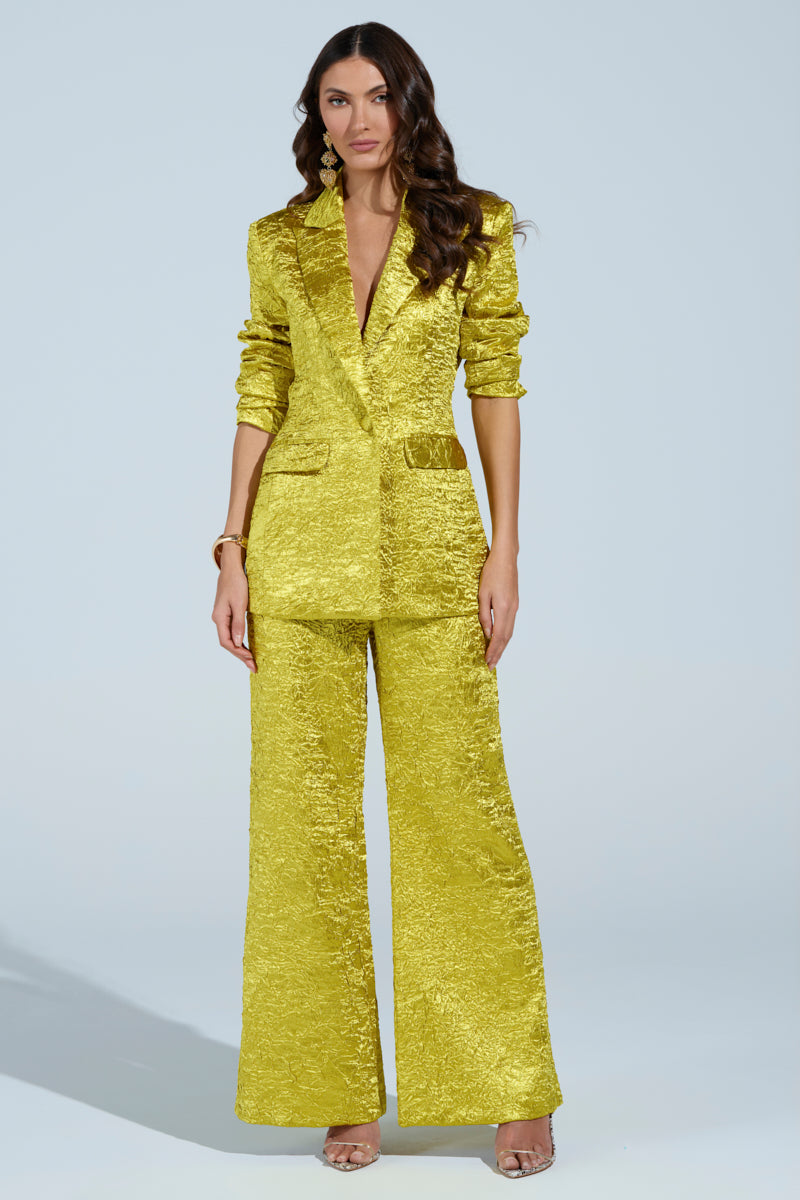 A woman in a textured, golden-yellow pantsuit from the AKIRA Label poses against a light gray background. Her long, wavy hair cascades elegantly as she dons matching earrings and the Expensive To Be Me Blazer, with her relaxed arms hanging gracefully by her sides.
