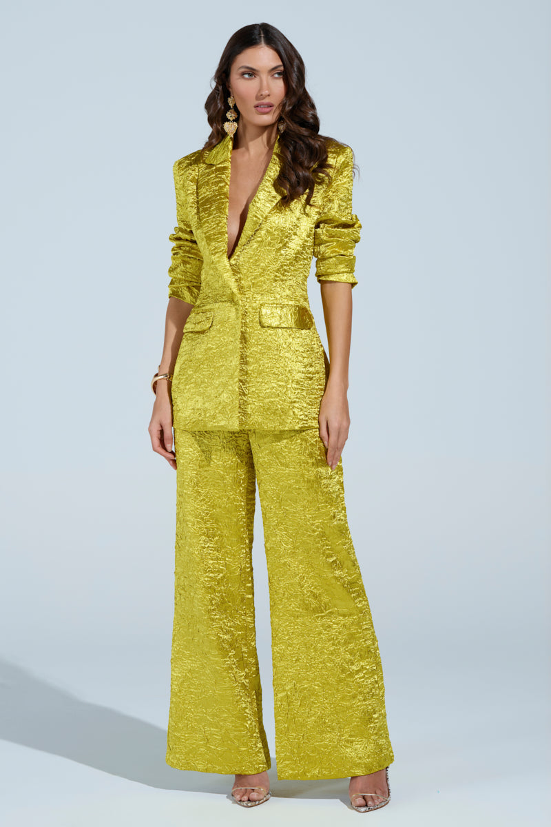 A woman confidently wears the AKIRA Label's vibrant yellow EXPENSIVE TO BE ME BLAZER pantsuit, her long wavy hair cascading over her shoulders. She accessorizes with statement earrings and a bracelet, pairing it all with neutral high-heeled sandals against a light blue backdrop.