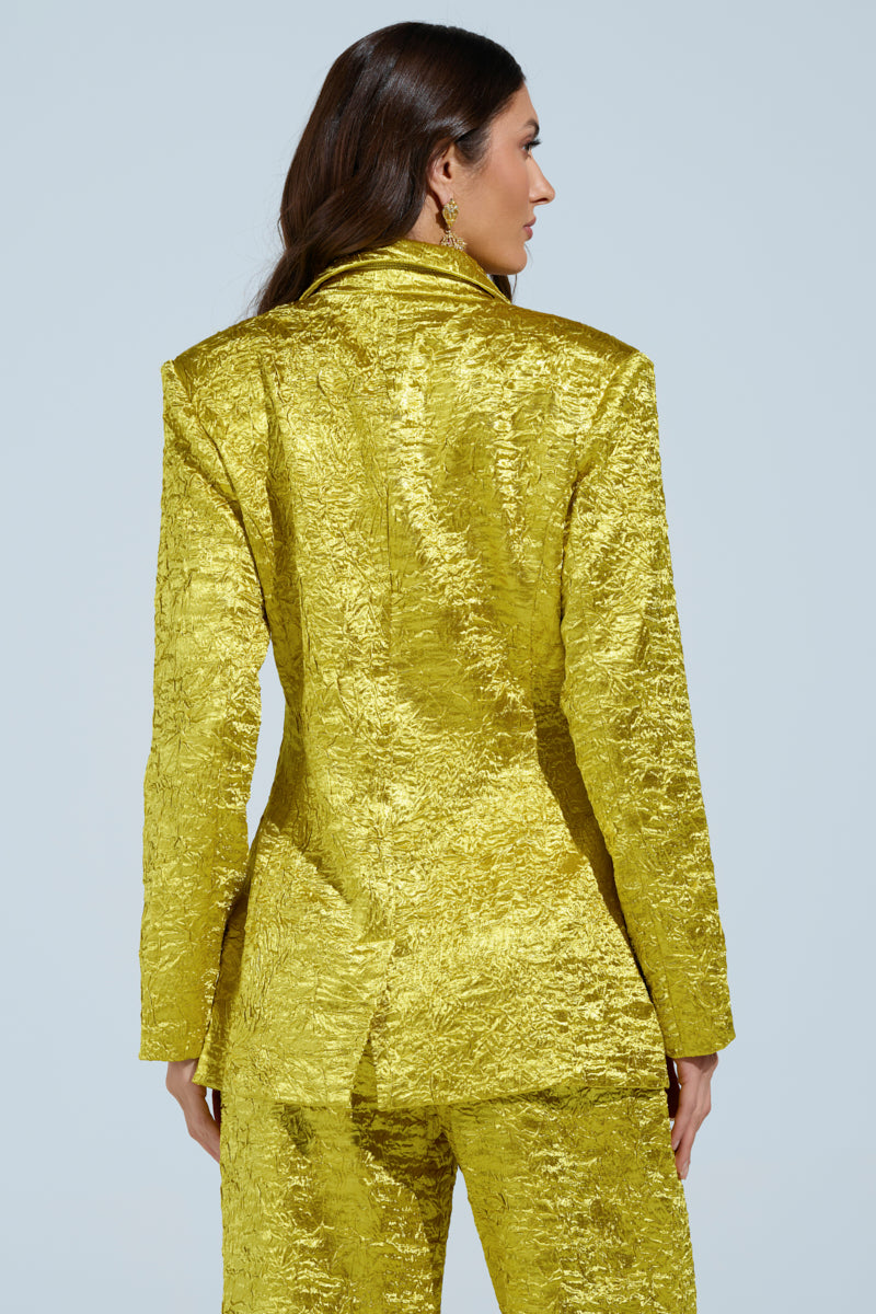 A person with long hair is facing away, wearing a shiny, crinkled gold AKIRA Label EXPENSIVE TO BE ME BLAZER and matching pants against a solid light blue background.
