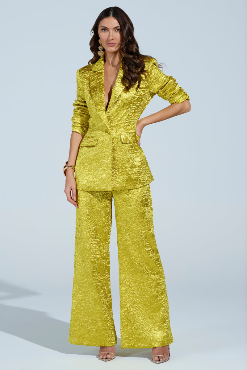 A woman stands confidently in a textured, bright yellow AKIRA Label pantsuit featuring wide-legged trousers and the "Expensive To Be Me Blazer." She accessorizes with gold earrings and bracelets, her long wavy hair complementing the plain, light-colored background.