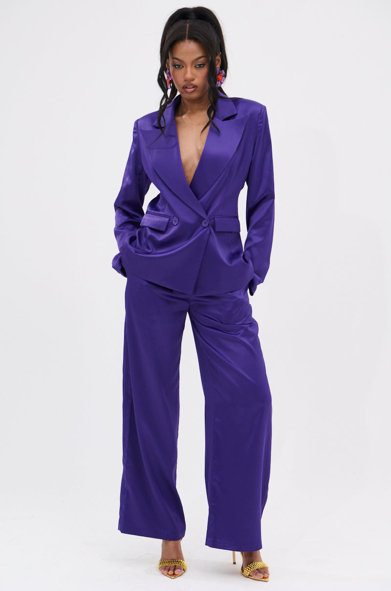 She models the SAY YOU LOVE ME SATIN TROUSER, a bold deep purple wide-leg pantsuit with a plunging neckline. Her long, dark hair falls in waves as she accessorizes with statement earrings and yellow high-heeled sandals.