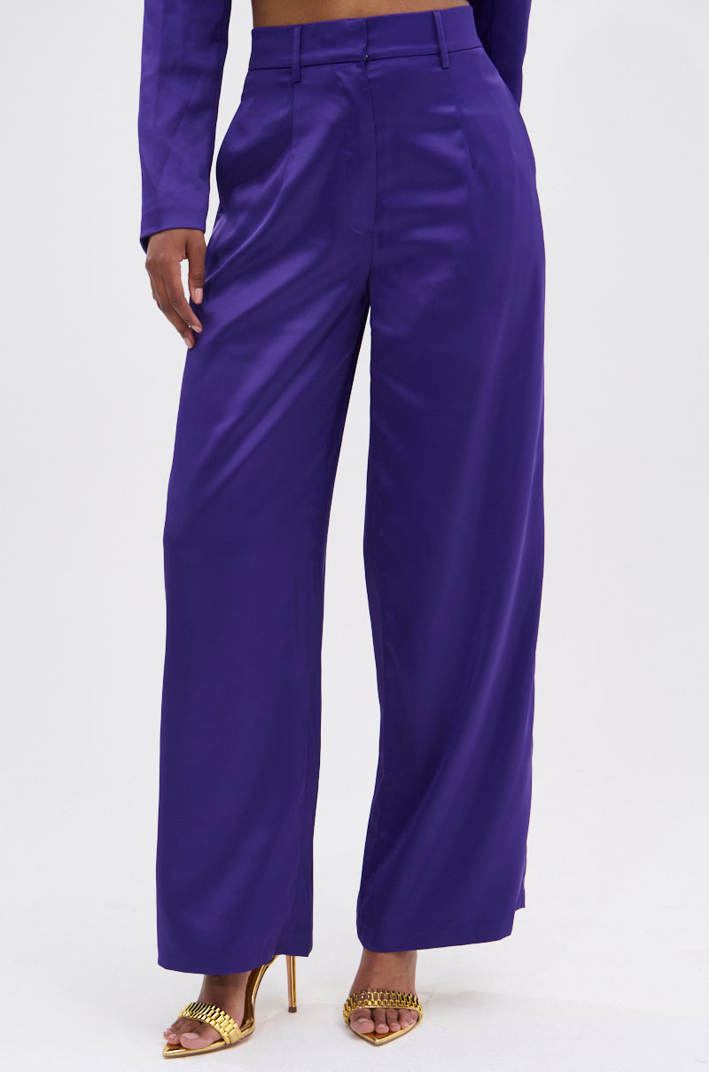 Wearing SAY YOU LOVE ME SATIN TROUSER in purple with a high-rise, wide-leg fit, gold open-toe high-heeled sandals, and part of a matching purple long-sleeve top visible.