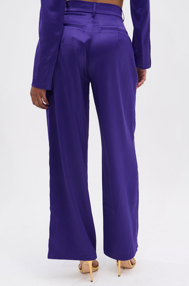 A person wearing the SAY YOU LOVE ME SATIN TROUSER in purple, featuring a high-rise fit and wide leg silhouette, stands with their back to the camera as tan heels peek out under the flowing pants paired with a matching jacket.