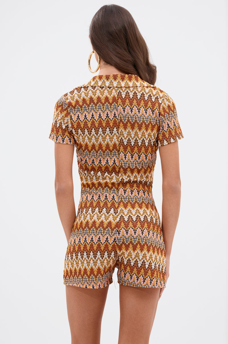 A woman with long brown hair, wearing large hoop earrings and the RYLEE ABSTRACT PRINT COLLARED ROMPER IN BROWN, stands facing away against a plain white background.