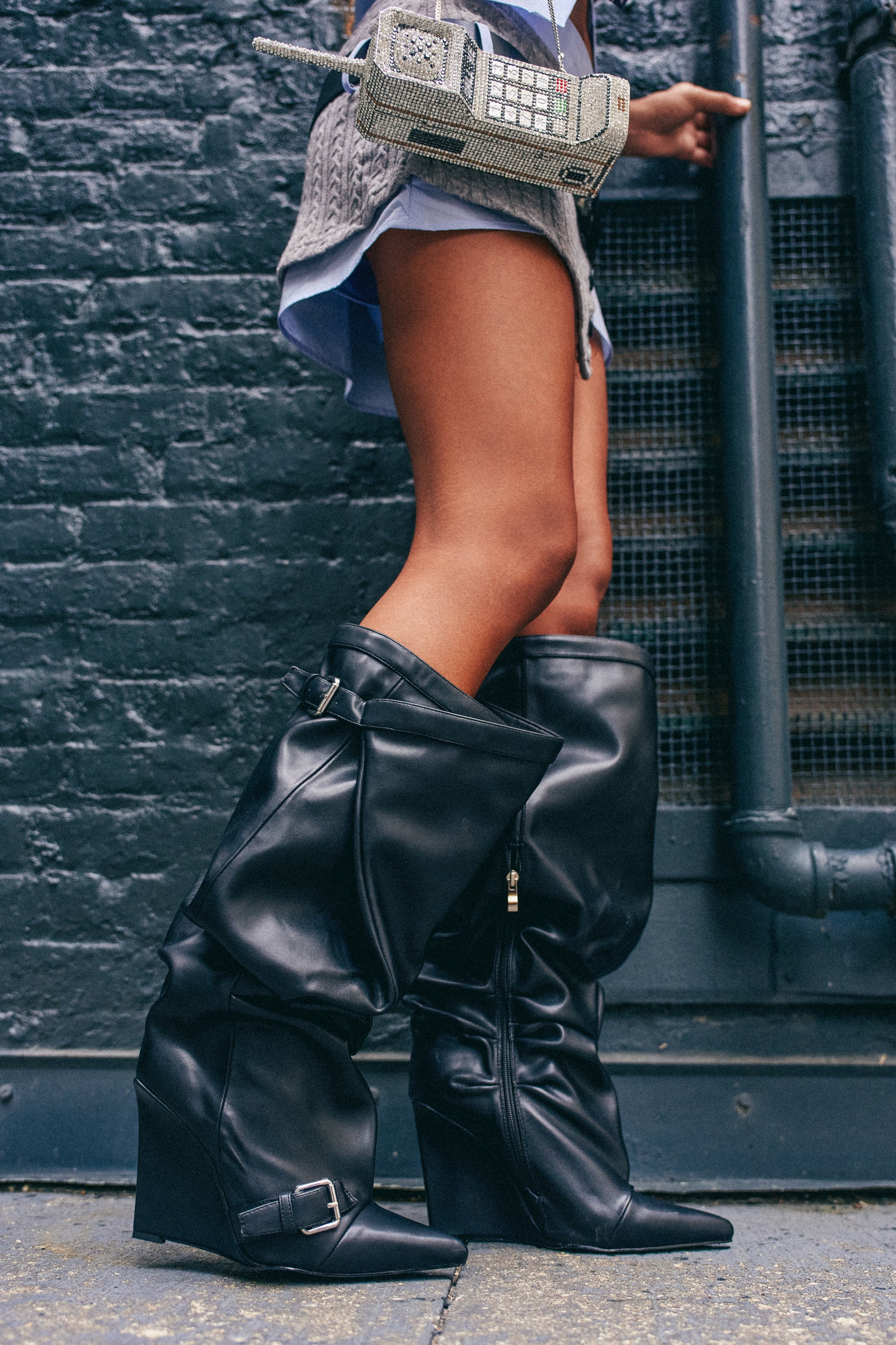 Wearing the AZALEA WANG ARACELI Black Belted Detail Wedge Boot and holding a small, retro cellphone-shaped purse, a person stands against a brick wall with exposed pipes.