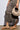 A person in a brown animal print skirt wears AKIRA ARCADIANA BROWN EMBELLISHED RHINESTONE SANDALS and holds a beige, textured round handbag. The faux leather accessories are modern and stylish, emphasizing earthy tones against a plain white background.
