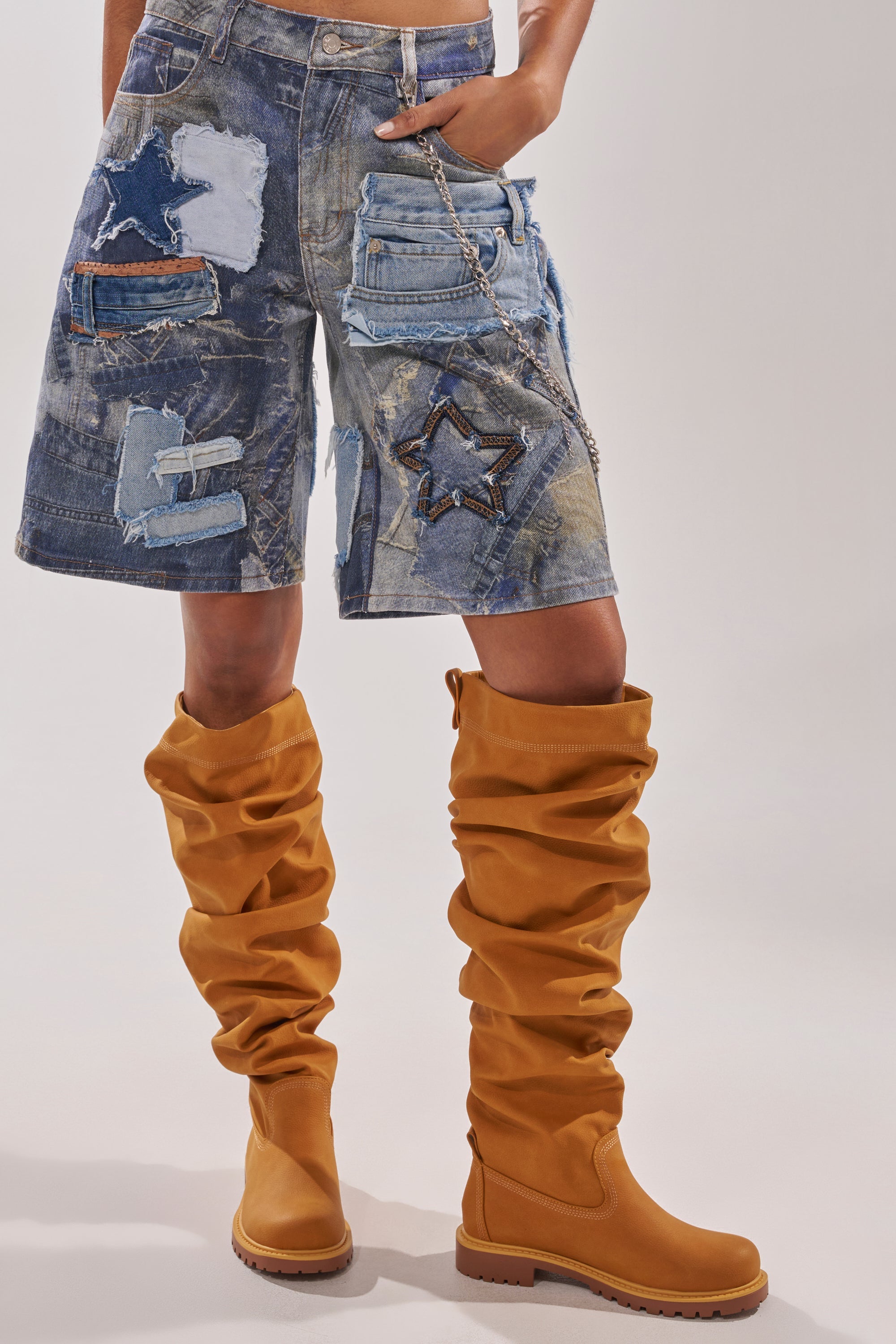 A person wearing distressed patchwork denim shorts with star-shaped patches and chains, paired with AZALEA WANG ARDIS TAN RUCHED FLAT BOOTS. The upper body is not visible. 2