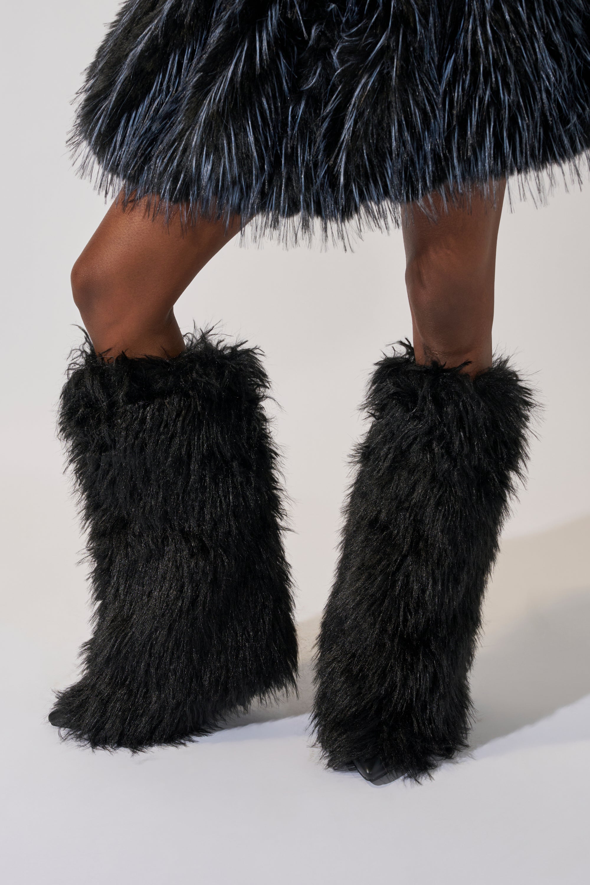 Only the lower half of a person wearing the AZALEA WANG ARETE BLACK FUR STILETTO BOOT and a matching black furry skirt is visible as they stand against a plain light background.