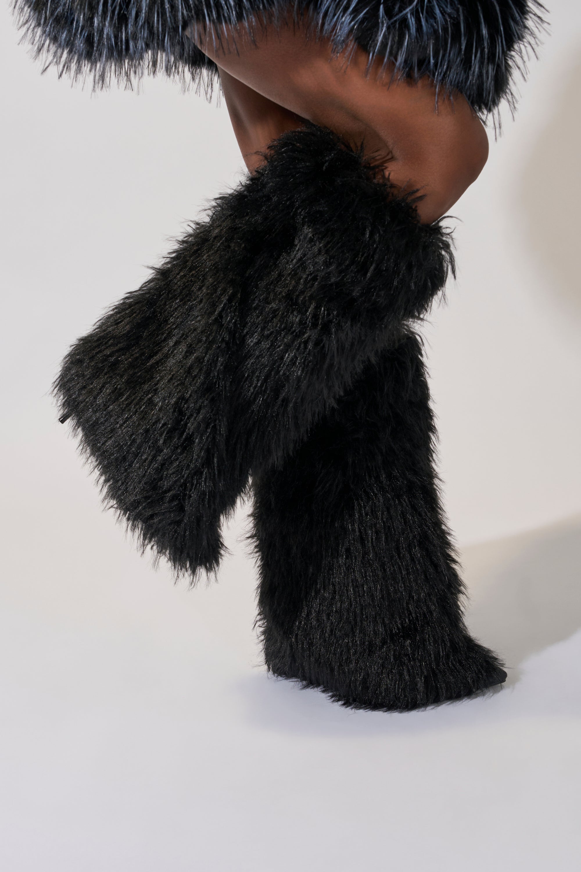A person stands with legs crossed against a plain light gray background, wearing the AZALEA WANG ARETE BLACK FUR STILETTO BOOT featuring black fur and a shaggy texture.