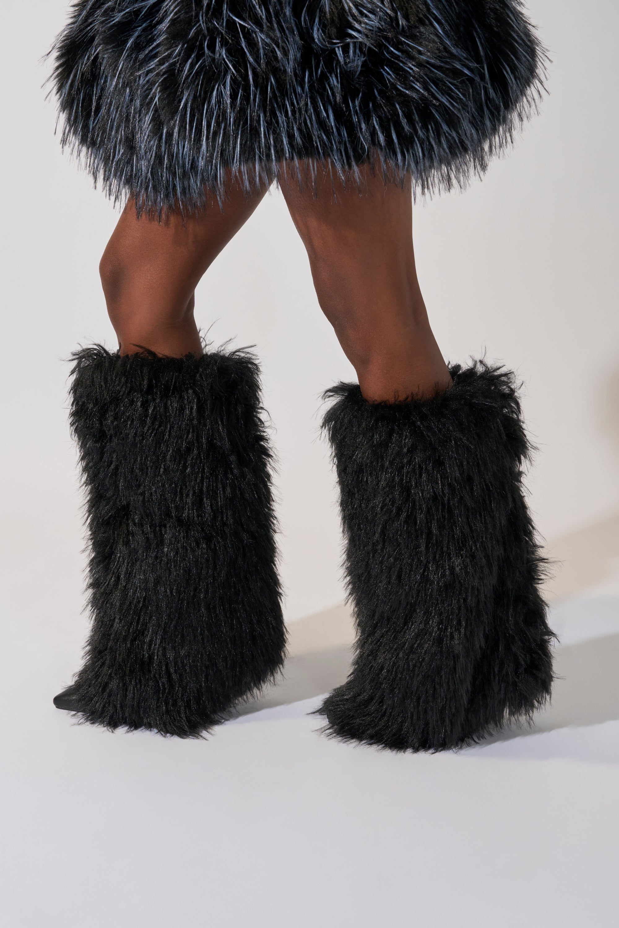 A model stands against a plain background, legs shown, wearing the AZALEA WANG ARETE BLACK FUR STILETTO BOOT paired with a matching black furry skirt.