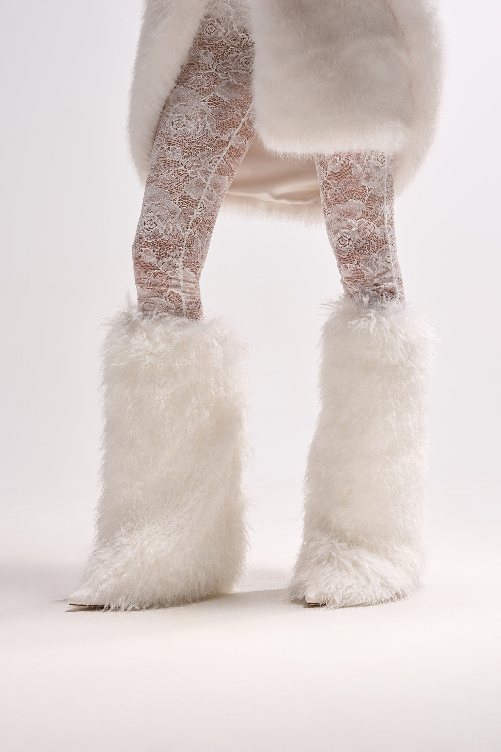 A person wearing white lace tights and the AZALEA WANG ARETE WHITE FUR STILETTO BOOT stands against a plain white background. Only the lower half, partly covered by a white faux fur coat, is visible.