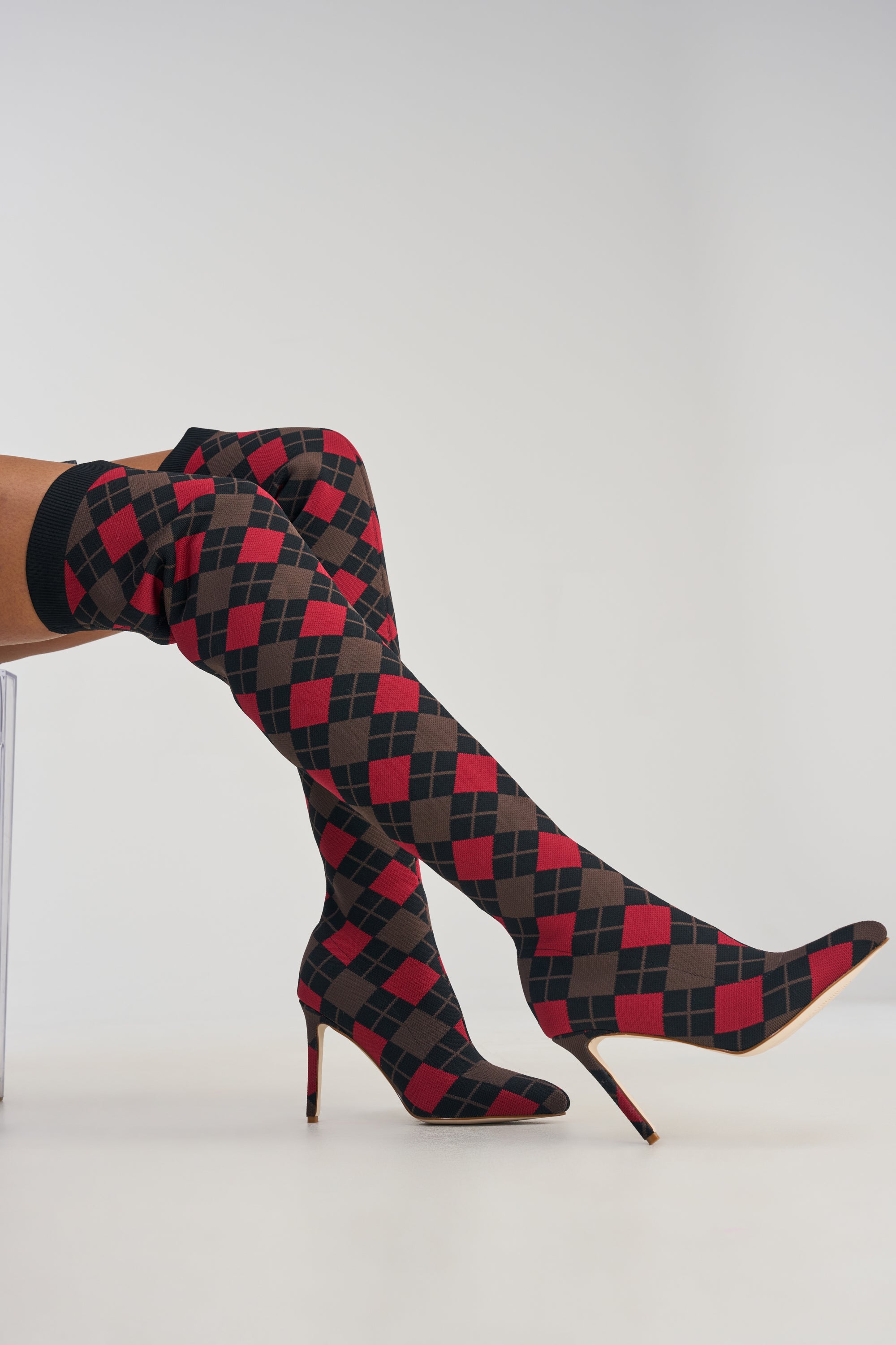A person wearing the AZALEA WANG ARGYLE BROWN MULTI STRETCH STILETTO BOOT, featuring an over-the-knee design with a bold red and black diamond pattern, sits with legs crossed against a plain light background.